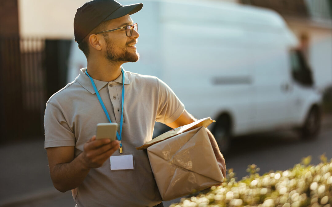 Happy delivery man text messaging on mobile phone while carrying packages.